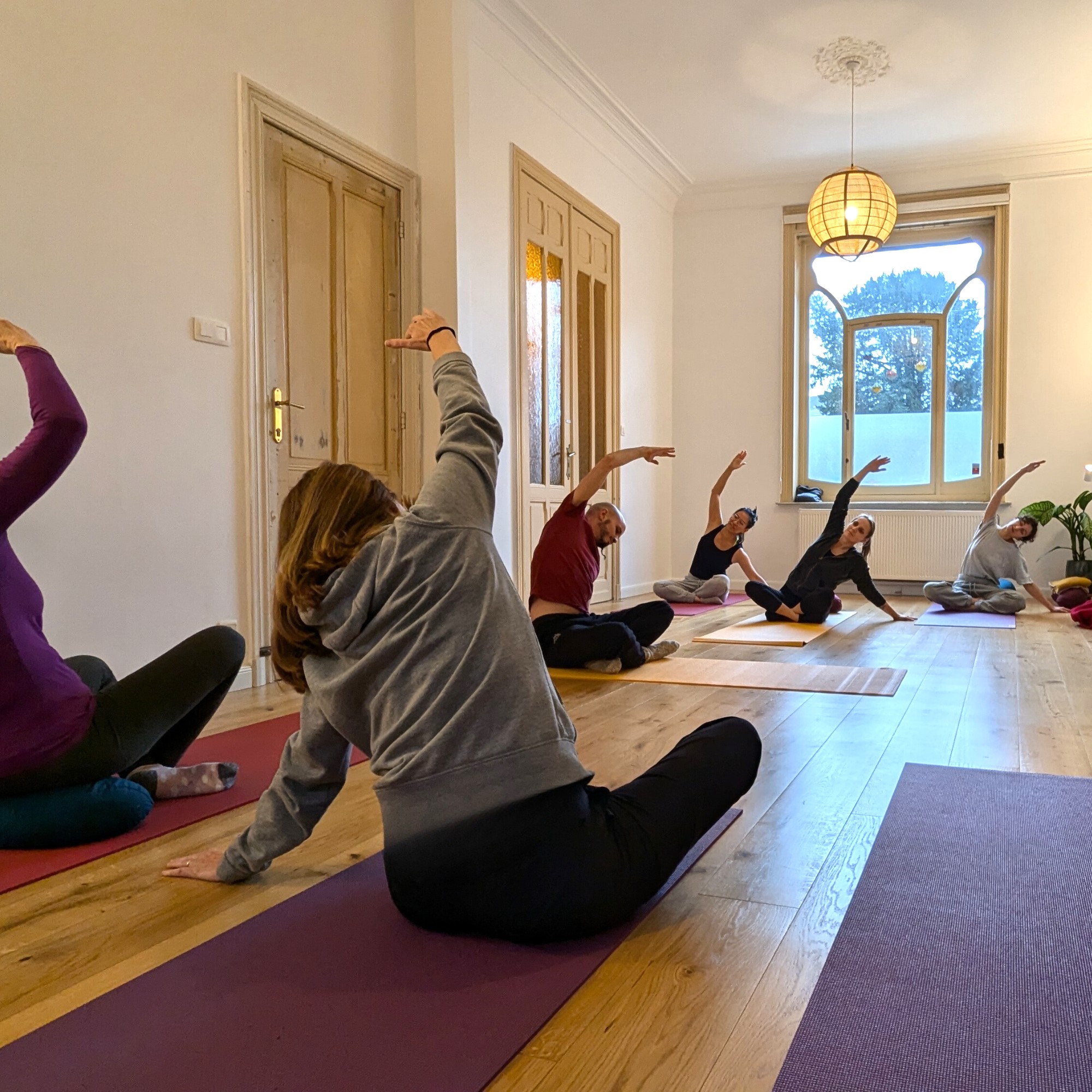 Salle de Yoga avec gens assis en tailleurs étirant le côté de leur dos.